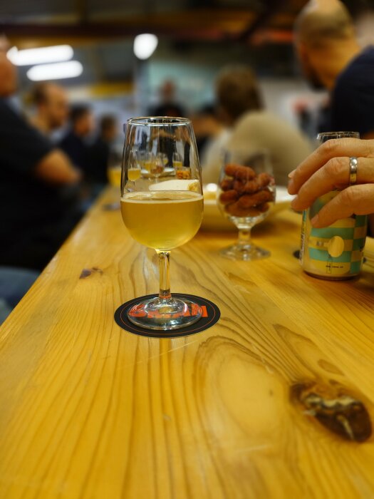 Ett glas öl står på ett träbord under en bryggerivisning med en grupp människor i bakgrunden.