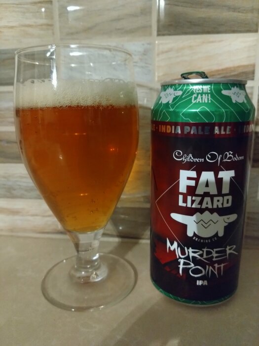 Glas med bärnstensfärgad IPA och en burk Fat Lizard Murder Point öl på beige bänkskiva.