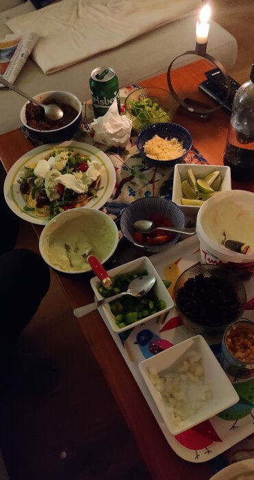 Tacobuffé på bord med grönsaker, toppings, en taccofylld tallrik, och en ölburk under tända ljus.