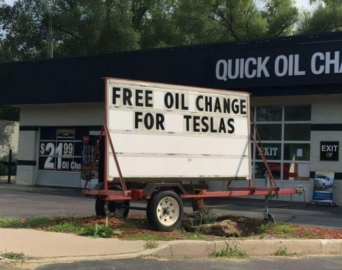 Skylt framför bilverkstad med texten "FREE OIL CHANGE FOR TESLAS" bredvid en oljebyteskampanj för $21.99.