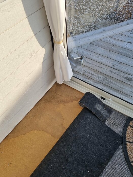 Fuktskada under golvmatta vid glasdörr i uterum med trätrall utanför.