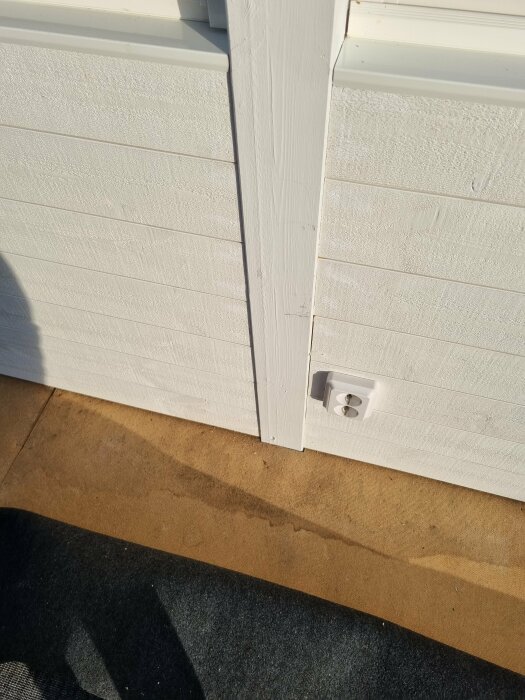 Vit träpanel med vägguttag nära golv, brun och svart matta synlig i förgrunden.