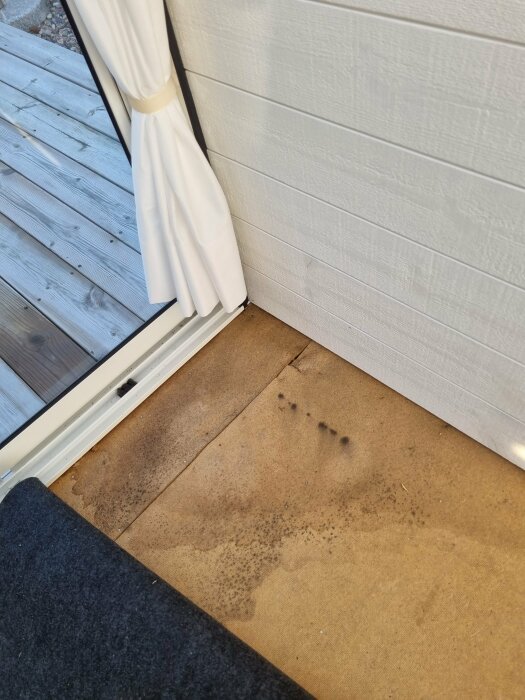 Fuktskadade golvbrädor vid en altandörr med vit gardin, bredvid en vägg av träpanel.