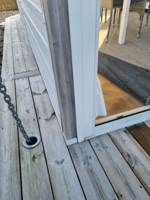 Närbild av hörn på veranda med trätrall och glasdörr, kedja fastsatt i golvet, med insyn till matplats.