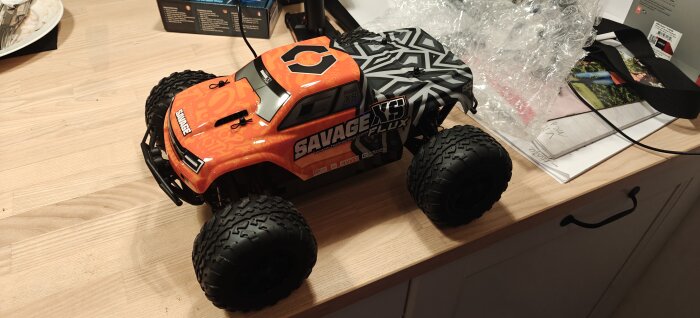 Orange RC-bil med stora däck och texten "SAVAGE" på karossen, placerad på ett träbord.