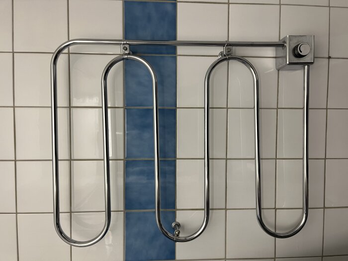 Kromad handdukstork med S-formad design, monterad på kaklad vägg, inklusive en kontrollenhet.