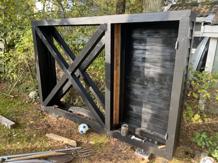 Svartmålat trästativ med snedställda balkar och skjuthakar, ståendes utomhus. Bakom ligger verktyg och en fotboll på marken.