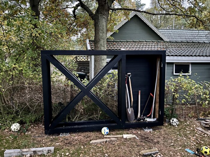 Svart träram till skjutregel bredvid grönt hus, omgiven av träd och buskar, med trädgårdsredskap och fotbollar på marken.