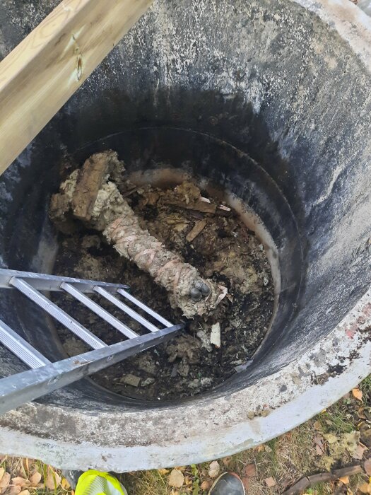 Öppen brunn med trasig isolering runt en vattenstam, stege ned i brunnen, omgiven av mark och löv.