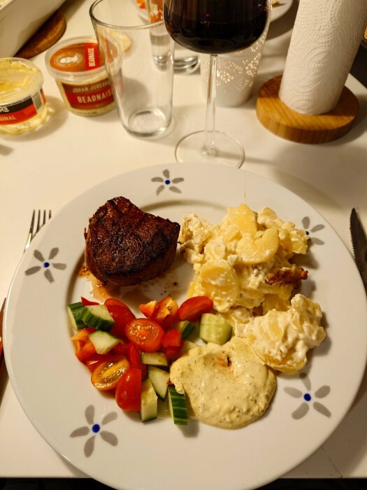 Grillad rostas med potatisgratäng, grönsaker och bearnaisesås på en tallrik, med ett glas rött vin i bakgrunden.