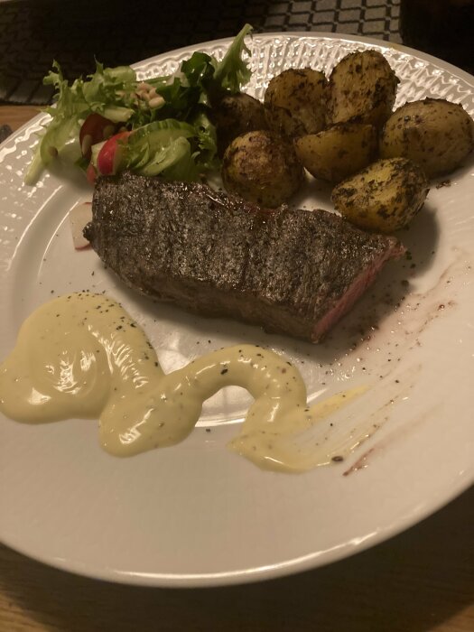 Grillad ryggbiff med grillade potatisar, grönsallad med rostade pinjenötter och sås på en tallrik.