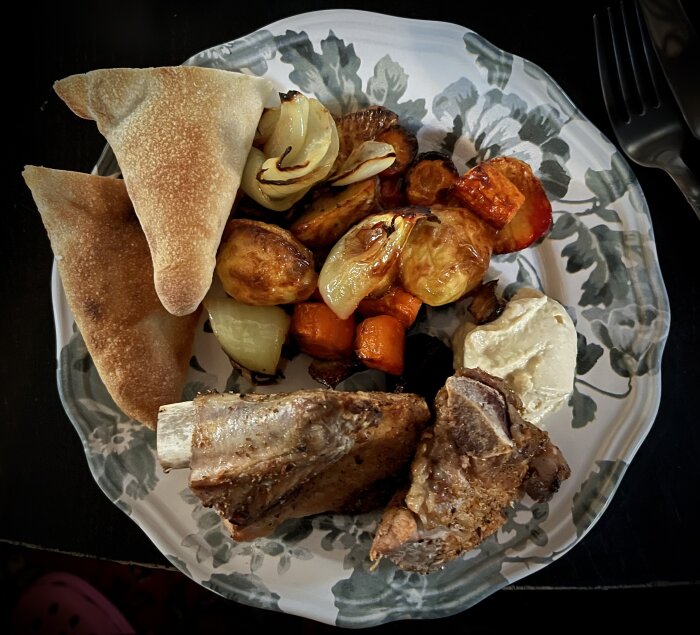 Tallrik med grillade revben, ugnsrostade grönsaker, bröd och hummus.