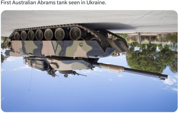 Stridsvagn i kamouflagemönster på betongväg, omgiven av träd och blå himmel i bakgrunden.