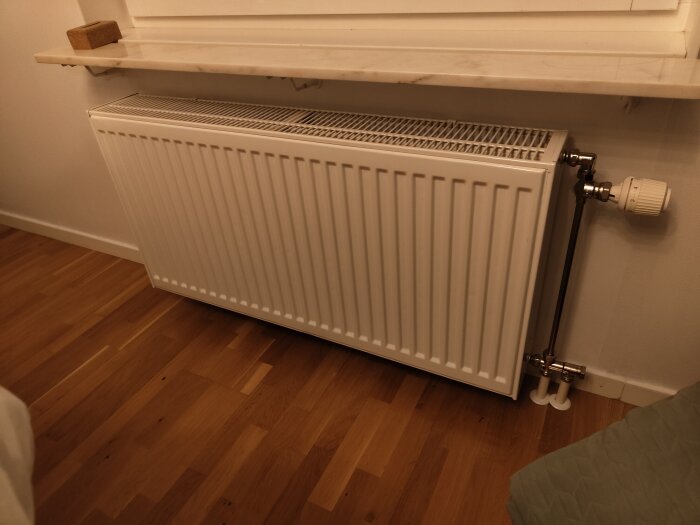 Vit radiator med termostat placerad i ett rum med parkettgolv och ett fönsterbräde ovanför.