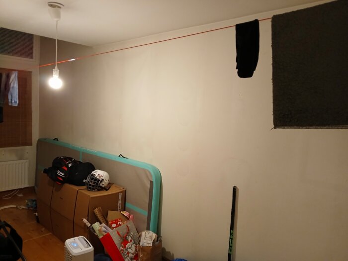 Rummet med gipsskivor, en glödlampa hänger från taket, kartonger och sportutrustning på golvet, förberett för renovering och isolering.