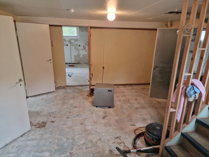 Källargolv i gammalt hus med slitna plattor, flera öppna dörrar, städutrustning och en lampande taklampa. Rör väntar på renovering.