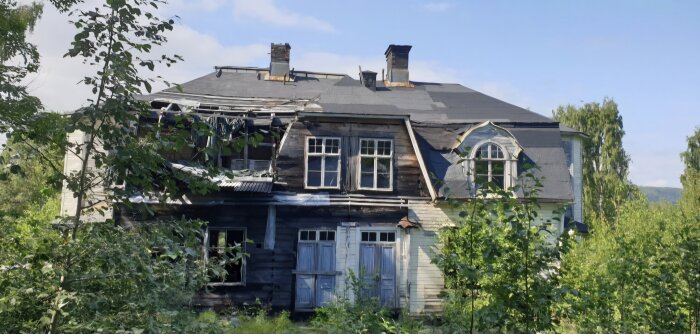 Övergivet hus med trasigt tak och fönster, delvis skymt av träd och växtlighet, beläget i en skogsmiljö under en klar himmel.