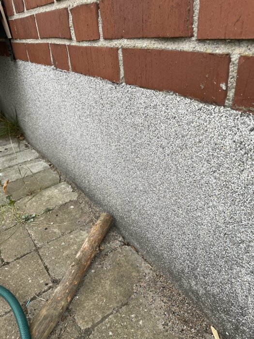 Bild på grund och fasad med röd tegelvägg ovanpå, nära betongyta och tegelstenar, med trästock och slang på marken.