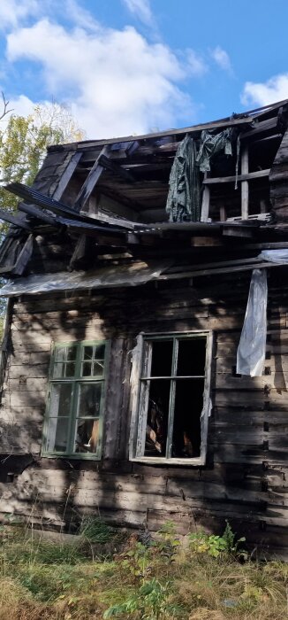 Förfallet trähus med trasigt tak, slitna väggar och krossade fönster, omgiven av vildvuxet gräs och träd under blå himmel.