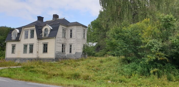 Gammalt, vitt trähus med svart tak och slitna fasader står vid en grönskande trädgård och skog.