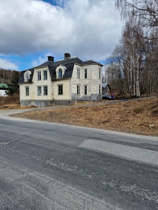 Gammalt tvåvåningshus med trasigt tak i landsbygdsmiljö, omgiven av bar skog och tom väg under molnig himmel.