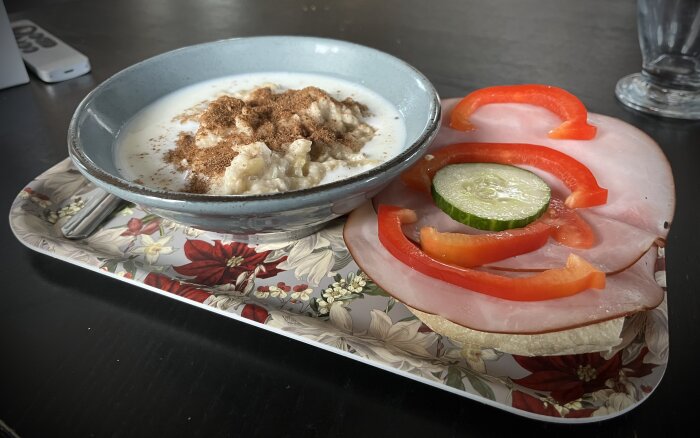 En skål med havregrynsgröt toppad med kanel och mjölk bredvid en skinkmacka med gurka och paprika på en blommig bricka.