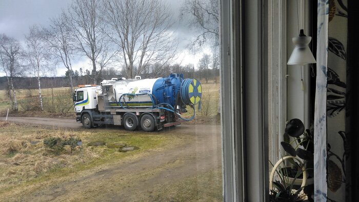 Slambil står parkerad utanför ett hus på landsbygden, fotograferad från insidan genom ett fönster.