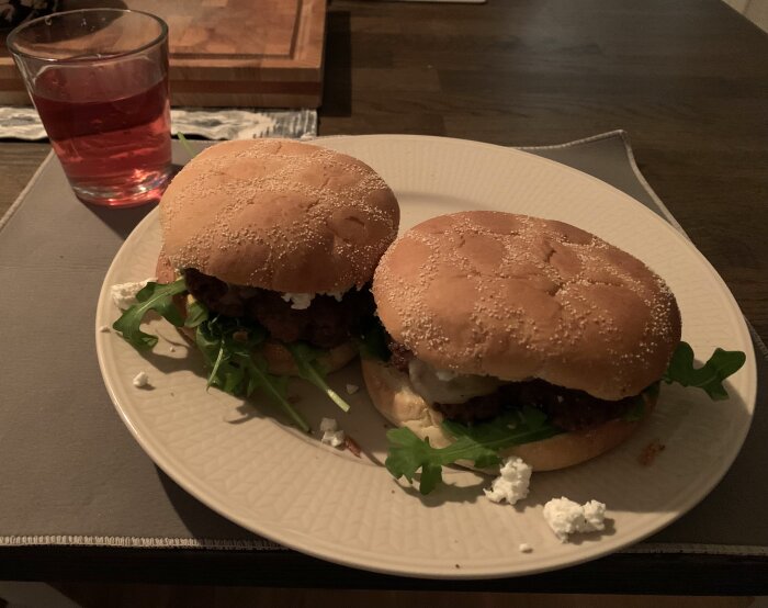 Två hamburgare på lammfärs med ruccola, feta, rödlök, ost, Srirachamajonäs och rostad lök på en tallrik, med ett glas dryck bredvid.