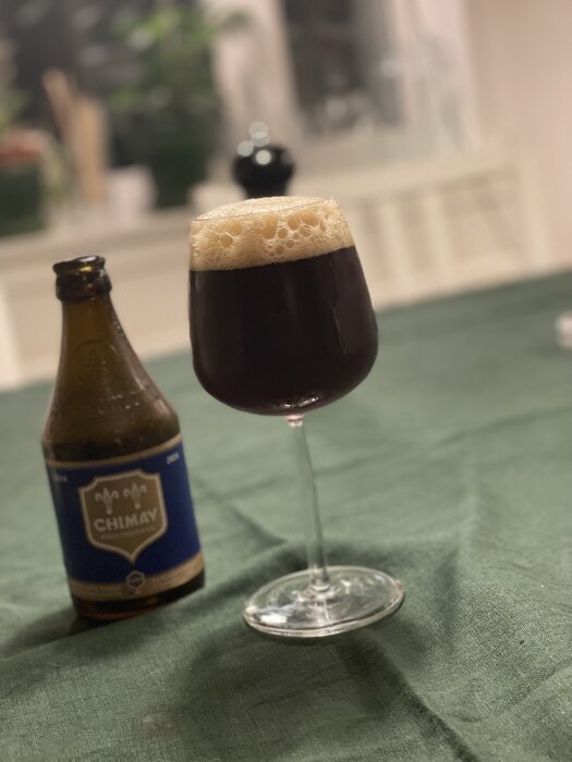 Mörkt öl i vinglas och en Chimay-flaska på grön duk, inomhusmiljö med suddig bakgrund.