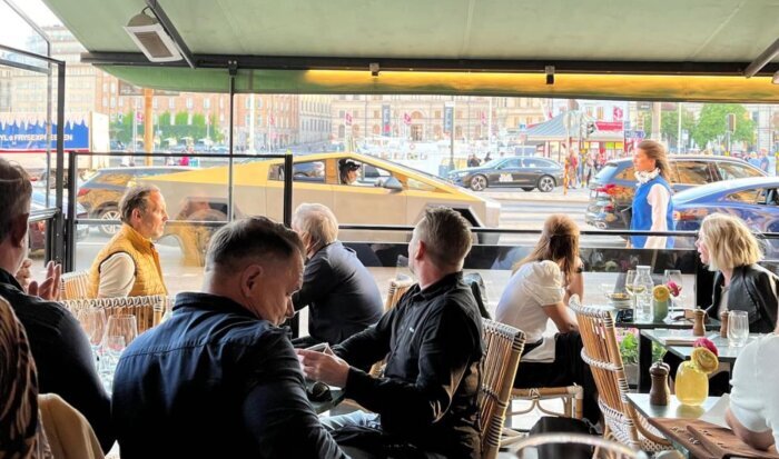 Restauranggäster sitter vid bord med en futuristisk bil som passerar utanför fönstret.