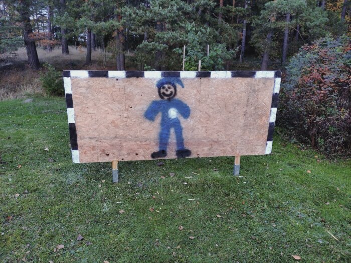 Måltavla gjord av spånskiva med målade svarta och vita ränder samt en blå streckgubbe; placerad utomhus på gräs med skog i bakgrunden.