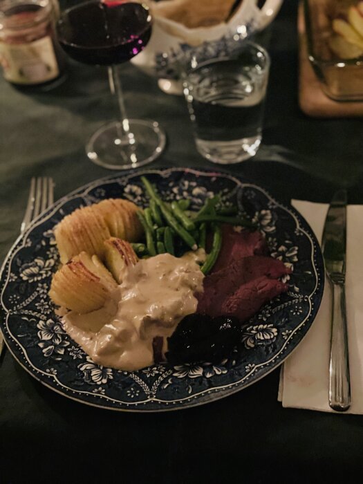 Tallrik med älgstek, potatis, grönsaker och sås, serverad på mönstrad porslinstallrik; rödvinsglas och vattenglas i bakgrunden.