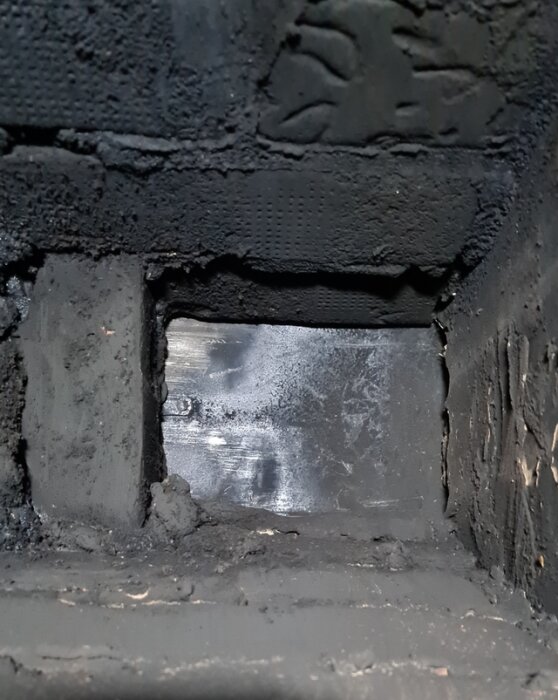 Närbild av ett sotigt ventilationsspjäll i en murstock, spjället syns vara delvis blockerat med sot och smågrus, vilket hindrar rörelse.