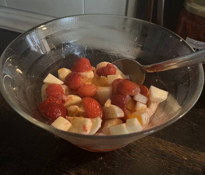 Glas-skål med fruktsallad; jordgubbar, melonbitar och en sked, serveras som tillbehör till risgrynsplättar.