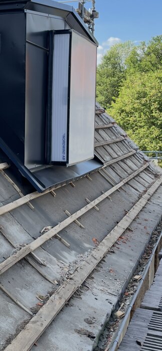 Tak under rivning med gammal beklädnad borttagen, visar träreglar och underlag; en ventilationsanordning syns på taket.