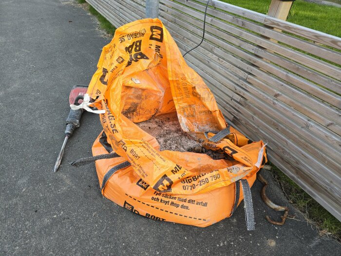 Öppen orange storsäck fylld med härdad betong placerad vid en trävägg, med en betongborr liggandes bredvid på asfalten.