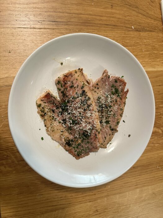 Tallrik med stekt fiskfilé, kryddad med örter och riven ost, serverad på ett träbord.