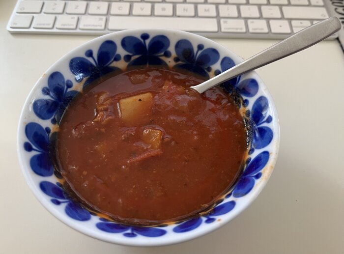 En skål med gulaschsoppa, serverad i en mönstrad blåvit skål med en sked i, ståendes på ett bord framför ett tangentbord.