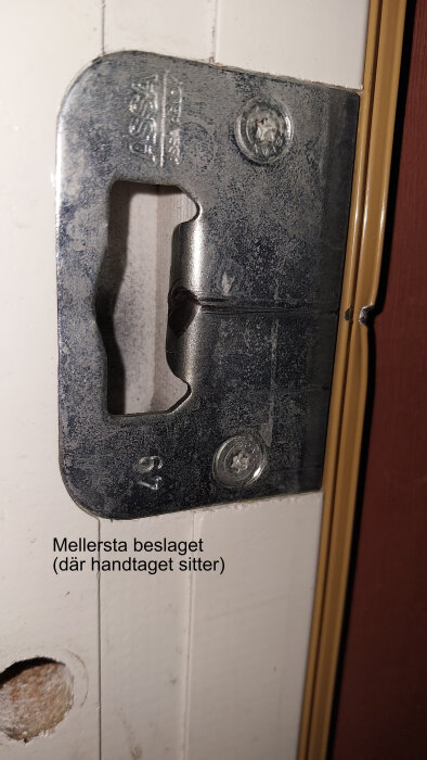 Slitet metallbeslag på dörrkarm, märkt "ASSA", med skruvhål och text "Mellersta beslaget (där handtaget sitter)" synligt.