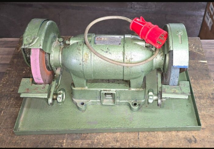 Grön slipmaskin med två slipstenar på arbetsbänk, ansluten med röd elkontakt.