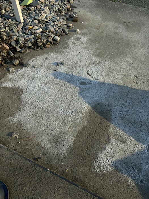 Betongplatta med vita fläckar, omgiven av små stenar. Möjliga fuktskador eller mögel på garageplatta. Skugga syns i vänstra hörnet.