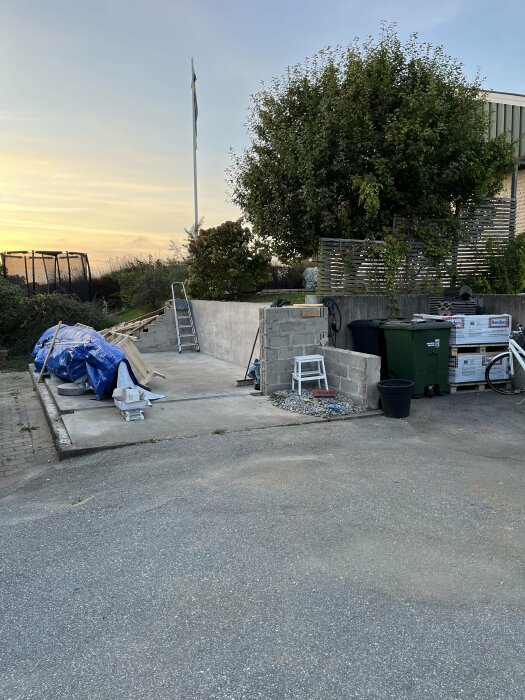 Betongplatta från garage med byggmaterial och byggavfall, omgiven av träd och staket, en stege lutar mot väggen under en klar himmel.