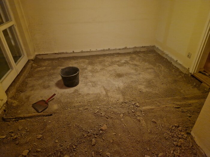 Rum med borttaget golv, hink och spade på ett lager sand under renovering.