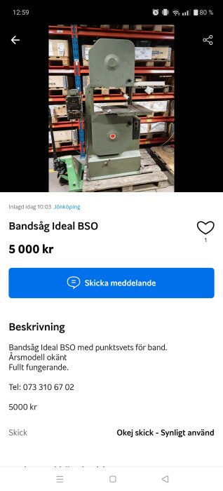 Grön bandsåg på en träpall i lagerutrymme, omgiven av hyllor med förpackningar.