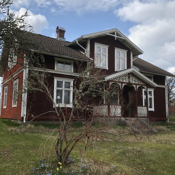 Ett rött trähus från tidigt 1900-tal med vita detaljer och verandaingång, omgiven av träd och gräsmatta under blå himmel.