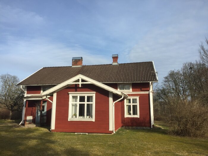 Rött trähus med vita knutar från tidigt 1900-tal på landsbygden. Huset har två skorstenar och ligger i en sluttning, omgiven av gräs och träd.