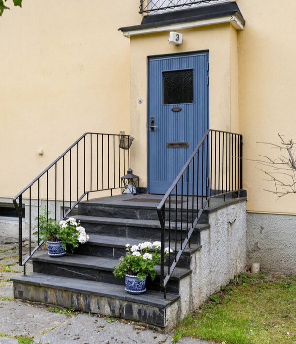 Blå dörr med svart räcke och nyrenoverad entrétrappa med blommor i krukor på sidan av ett hus med gul fasad.