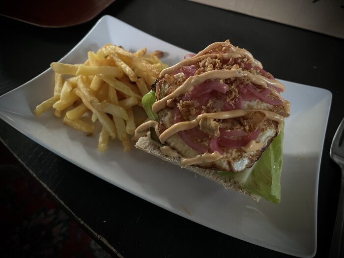 Hemmagjord hamburgare på ciabatta med sallad, stekt ägg, picklad och rostad lök, dressing, samt pommes frites på en vit tallrik.