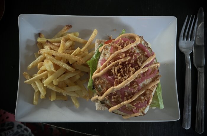 Hamburgare på ciabatta med stekt ägg, picklad och rostad lök, sallad, senap, ketchup och dressing. Serverad med pommes frites på en tallrik.