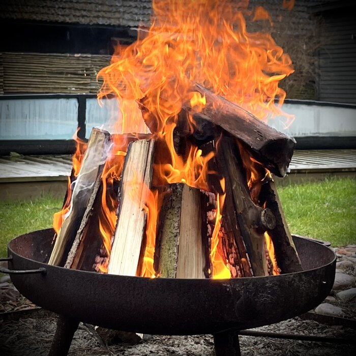 Brasa i eldfat med vedträn av gammal fur, omgiven av grön gräsmatta och träterrass i bakgrunden.